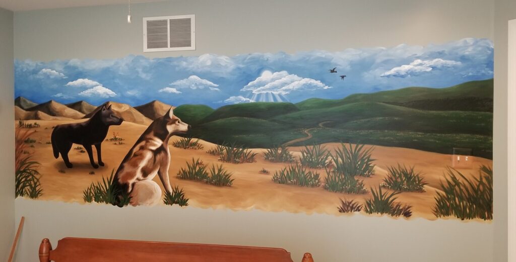 A gray wall painted from end to end with a mural of a landscape depicted a desert foreground with both mountains and grassy hills in the background. Two wolves sit on a sandy rock in the foreground, a black one and a brown and white one. Both wolves look to the right over the whole landscape.